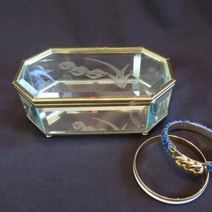 Puede incluir: Una caja de joyería de vidrio con un borde dorado y un diseño floral grabado. La caja está sobre una superficie negra con dos pulseras azules y doradas.