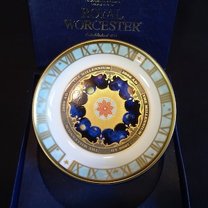 Peut inclure: Assiette commémorative Royal Worcester pour le Millénaire, présentée dans un coffret bleu. L'assiette présente un bord doré, des chiffres romains et un motif central avec des planètes et le texte "2000 AD THE MILLENNIUM".