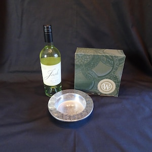 May include: A bottle of Josh wine next to a decorative silver dish with a patterned rim and a green box with a circular logo. The wine bottle is clear with a white label. The box is green with a decorative pattern.