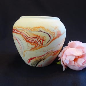 May include: Cream-coloured ceramic vase with a marbled design in shades of red, orange, and brown. The vase has a rounded shape and a wide opening. A pink rose is next to the vase.