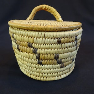 Può includere: Un piccolo cesto rotondo intrecciato con un coperchio e un manico. Il cesto è fatto di materiali naturali e ha un motivo marrone e beige.