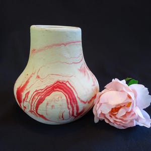 May include: A decorative vase with a marbled design in red and white. The vase has a rounded body and a flared opening. A pink rose with green leaves sits next to the vase against a dark background.