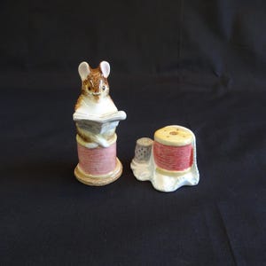 May include: A pair of ceramic figurines featuring a mouse reading a book atop a spool of thread and a spool with a thimble. The figurines are painted in shades of brown, pink, and cream, with fine detailing.