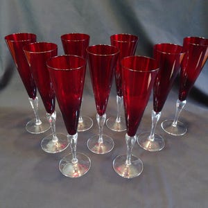 Peut inclure: Un ensemble de neuf flûtes à champagne hautes dotées de bols en verre rouge profond et de pieds en verre transparent avec des bases circulaires. Les verres sont disposés en triangle, mettant en valeur la couleur cramoisie et le design effilé.