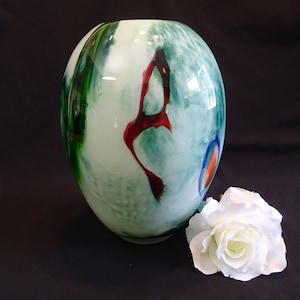 May include: A white glass vase with green, red, and blue abstract designs. The vase is sitting on a black surface with a white rose.