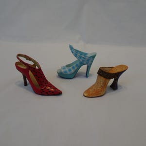May include: Three miniature high-heeled shoes in red, blue, and orange. The red shoe has a pointed toe and a slingback strap. The blue shoe has a gingham pattern and a peep-toe design. The orange shoe has a floral pattern.