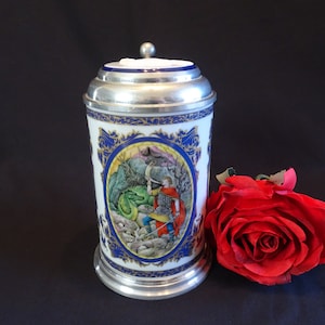 May include: A white ceramic beer stein with a silver lid and base. The stein features a colorful illustration of a knight battling a dragon. Blue and gold decorative trim surrounds the central image. A red rose rests beside the stein.