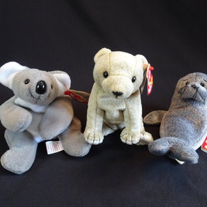 May include: Three plush stuffed animals: a gray koala, a beige polar bear, and a gray seal. Each animal has a red and white Ty tag attached.