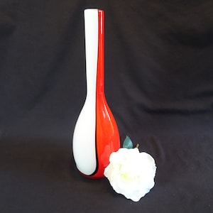 May include: A tall, white and red glass vase with a curved design. The vase is on a black background.
