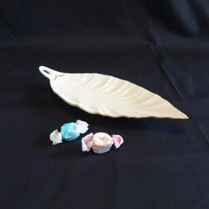 Può includere: Un piatto da portata a forma di foglia color crema con un bordo dorato. Due caramelle incartate, una blu e una rosa, sono accanto al piatto. Il piatto ha un manico e una superficie strutturata.