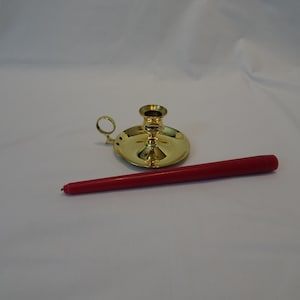 May include: A gold-colored candlestick holder with a round base and a small handle. A long, red taper candle rests beside the holder. The candle is cylindrical and tapers to a point. The items are on a white surface.
