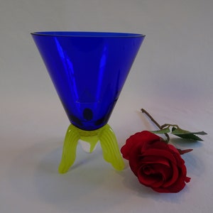 May include: A cobalt blue glass vase with a flared top and three yellow glass legs. A single red rose is lying on the white surface next to the vase.