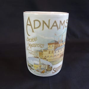 Puede incluir: Taza de cerámica blanca con un borde marrón. La taza presenta una ilustración vintage de una cervecería con el texto "Adnams Brewers & Maltsters Wine & Spirits Importers Suffolk" en letras doradas.