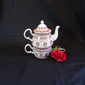 Könnte beinhalten: Ein weiß-rotes Keramik-Teeservice mit floralem Muster, bestehend aus einer Teekanne, einer Teetasse und einem Deckel. Eine rote Rose liegt neben dem Teeservice. Das Teeservice steht auf einer schwarzen Oberfläche.