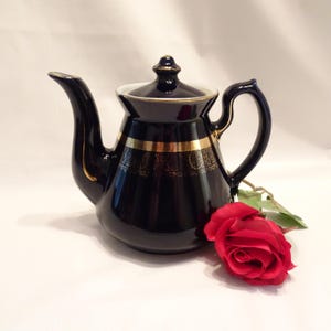 May include: A navy blue ceramic teapot with gold trim and a matching lid. The teapot has a curved spout and handle. A red rose rests beside the teapot on a white surface.