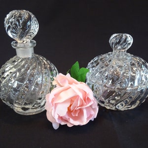 Puede incluir: Dos botellas de vidrio transparente con diseños de espiral texturizados. La botella de la izquierda es una botella de perfume con un tapón. La botella de la derecha es un frasco con una tapa. Una rosa rosa está delante de las botellas.