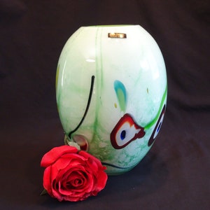 May include: A green glass vase with a white background and colorful abstract designs. The vase is round and has a smooth surface. A red rose is lying next to the vase.