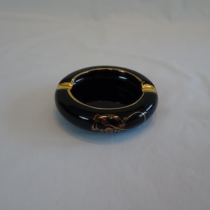 May include: A round, black ceramic ashtray featuring a glossy finish and gold-colored trim along the inner rim. The exterior surface is decorated with a detailed, circular graphic design in gold, red, and green tones, including two cigarette rests.