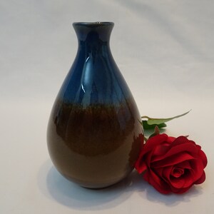 Puede incluir: Un pequeño jarrón de cerámica azul y marrón con cuerpo redondeado y cuello estrecho. Una sola rosa roja está tumbada junto al jarrón.