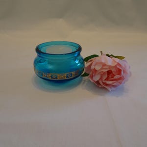 May include: A teal glass votive candle holder with a gold floral design around the middle. A white candle sits inside the holder. A pink rose with green leaves rests beside the candle holder.