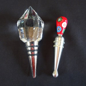 May include: Two bottle stoppers, one is a clear crystal with a faceted design and a silver base, the other is silver with a red and blue glass top with a floral pattern.