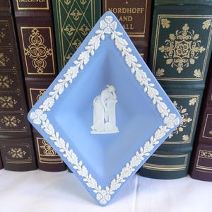 May include: A blue and white ceramic trinket dish with a diamond shape and a raised design of a woman leaning against a pillar. The dish has a white floral border.