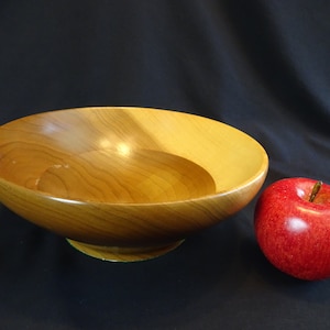 Puede incluir: Un tazón grande de madera con un acabado natural. El tazón es redondo y tiene una ligera curva en los lados. Está sentado sobre una superficie negra junto a una manzana roja.