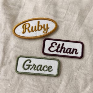 May include: Three embroidered fabric patches with white backgrounds and colored borders. One oval patch features the name Ruby in gold thread. Two rectangular patches feature the names Ethan in dark red and Grace in olive green thread.