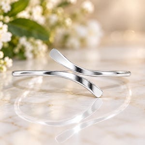 May include: A polished silver-tone bypass cuff bracelet featuring a smooth, open-ended design with two slightly curved, tapered ends that overlap slightly in the center, creating a sleek and reflective metallic appearance.