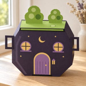 May include: A dark purple paper craft box shaped like a house with a light green roof, two green tree cutouts, a purple arched door, two yellow circular windows, a yellow crescent moon, and small yellow stars on the front surface.