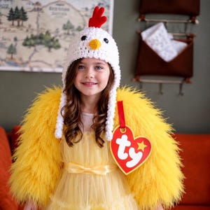 May include: A white crocheted chicken hat with a red crest and yellow beak, paired with a bright yellow feathered shrug and a large red heart-shaped tag featuring the white text "ty" and a yellow star, worn over a yellow dress.