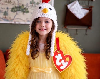 May include: A white crocheted chicken hat with a red crest and yellow beak, paired with a bright yellow feathered shrug and a large red heart-shaped tag featuring the white text "ty" and a yellow star, worn over a yellow dress.
