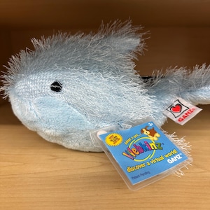 Puede incluir: Un peluche de color azul claro con pelaje desgreñado y un ojo negro. Incluye una etiqueta blanca con un corazón rojo y el texto GANZ, y una etiqueta de plástico azul que dice Webkinz, Adopt a pet, discover a virtual world, GANZ.