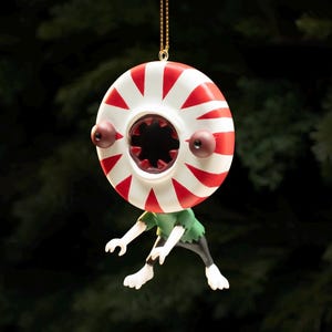 May include: A hanging ornament featuring a round, red and white striped peppermint candy head with two dark eyes and a central mouth filled with sharp teeth. The figure has a green shirt, black pants, and white limbs, suspended by a gold chain.