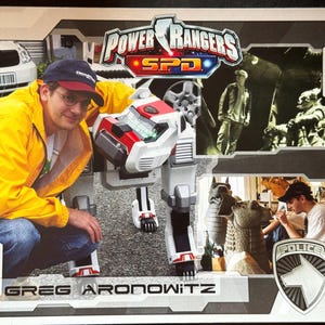 May include: A photograph featuring a person in a yellow jacket and a Power Rangers SPD robot. The image includes the text "Power Rangers SPD" and the name "Greg Aronowitz". Additional images are in the background.