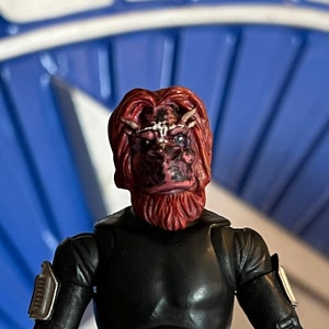 May include: A red-skinned action figure with a black and silver suit. The figure has a red beard and a detailed face with a white stripe across the forehead.