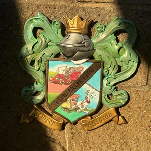Op de afbeelding: Een groen en gouden wapenschild met een kroon, een ridderhelm en een rode auto. Het schild heeft de woorden "Toad Hall" en "Accelerando" erop. De leuze is "Semper Absurda".