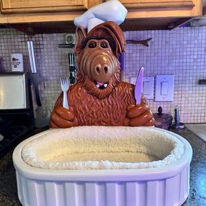 Puede incluir: Una cama para mascotas con forma de figura de chef de dibujos animados marrón, sosteniendo un tenedor y un cuchillo. La cama es blanca, rectangular y tiene un interior blanco y esponjoso.