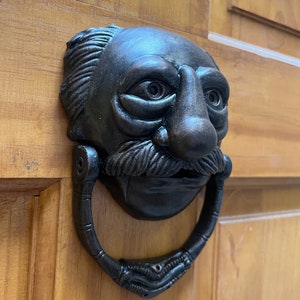 Puede incluir: Un llamador de puerta de metal negro con forma de cara de hombre con una nariz grande y un bigote. El llamador tiene un asa de anillo.