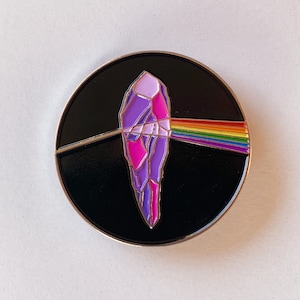 Puede incluir: Un pin de esmalte negro y plateado con un cristal morado y rosa con un rayo de prisma arcoíris que lo atraviesa.