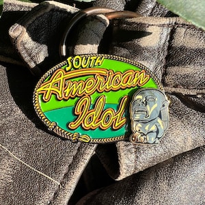 May include: A gold-colored enamel pin with a green background and the text "South American Idol" in yellow and orange lettering. A small, gray, enamel pin of a person's head is attached to the larger pin.