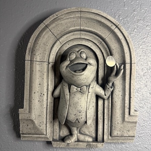 Puede incluir: Decoración de pared gris, similar a una piedra, con una rana de dibujos animados con esmoquin sosteniendo una moneda. La rana está enmarcada por un diseño de puerta arqueada. La escultura tiene un aspecto texturizado y moteado.