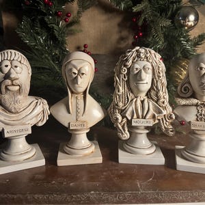 May include: Four off-white busts of historical figures: Aristotle, Dante, Moliere, and Shakespeare. Each bust is mounted on a square base with the name of the figure displayed on a small plaque. The busts are in front of a Christmas wreath.