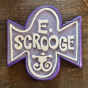 Puede incluir: Un letrero morado y blanco con el nombre "E. Scrooge" en letras estilizadas. El letrero tiene un fondo texturizado y un emblema decorativo en la parte inferior. El letrero está sobre una superficie de madera.