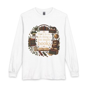 Op de afbeelding: Een wit shirt met lange mouwen en een centrale afbeelding van stapels boeken, een wereldbol, een bureaulamp, een mok en een slapende kat. De tekst "there is no such thing as too many books" staat in het midden, omringd door bladeren.