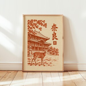 Könnte beinhalten: Ein gerahmter Kunstdruck auf hellbeigem Hintergrund mit einer rostfarbenen Illustration eines Hirsches vor einem traditionellen japanischen Tempelgebäude. Ahornblätter hängen darüber und die japanischen Schriftzeichen 奈良鹿 sind vertikal aufgedruckt.