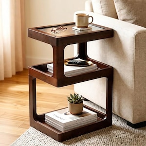May include: A dark brown wooden side table with three tiers. The top tier features a glass insert, a ceramic mug, and eyeglasses. The middle tier holds a remote control and books. The bottom tier holds a potted succulent and books.