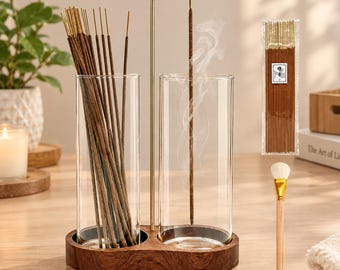 May include: A dual glass cylinder incense holder set on a dark wood base with a gold metal stand. One cylinder holds a bundle of brown incense sticks, while the other contains a single burning stick emitting smoke. Includes a small cleaning brush.