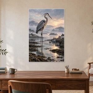 Op de afbeelding: Een verticale wandkunstprint met een grote grijze reiger die bij zonsopgang in ondiep kustwater staat. De achtergrond toont een mistige haven met kleine boten, rotsachtige oevers en verre heuvels onder een zachte, bewolkte lucht.