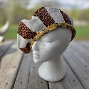 May include: A multi-paneled beret featuring alternating sections of solid off-white fabric and a brown and white geometric diamond pattern. The brim is trimmed with a gold-toned fabric band decorated with a repeating blue and gold emblem design.